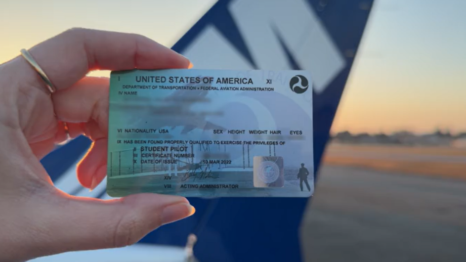 Female hand holding student pilot certificate in front of a blue Cessna 172 airplane tail at sunset