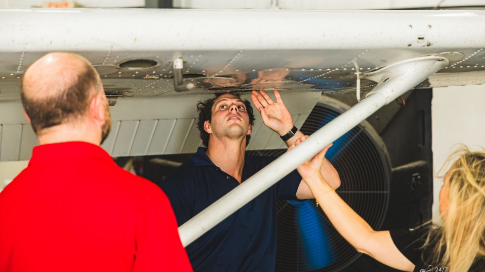 Two male certified flight instructors (CFIs) and a female student pilot inspecting a Cessna 172 airplane wing while in the maintenance hangar