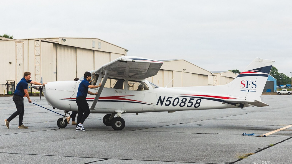 Two male student pilots pushing back a white Cessna 172 airplane with a tow bar