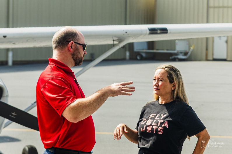 Certified flight instructor (CFI) in red shirt teaching female student pilot, wearing a Fly Delta Jets shirt, in front of Cessna 172 airplane wing